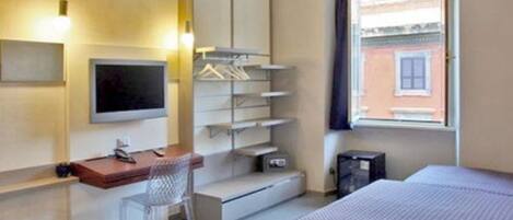 Quarto de hotel com cama, escrivaninha, cadeira, televisão e janela com vista para um edifício.