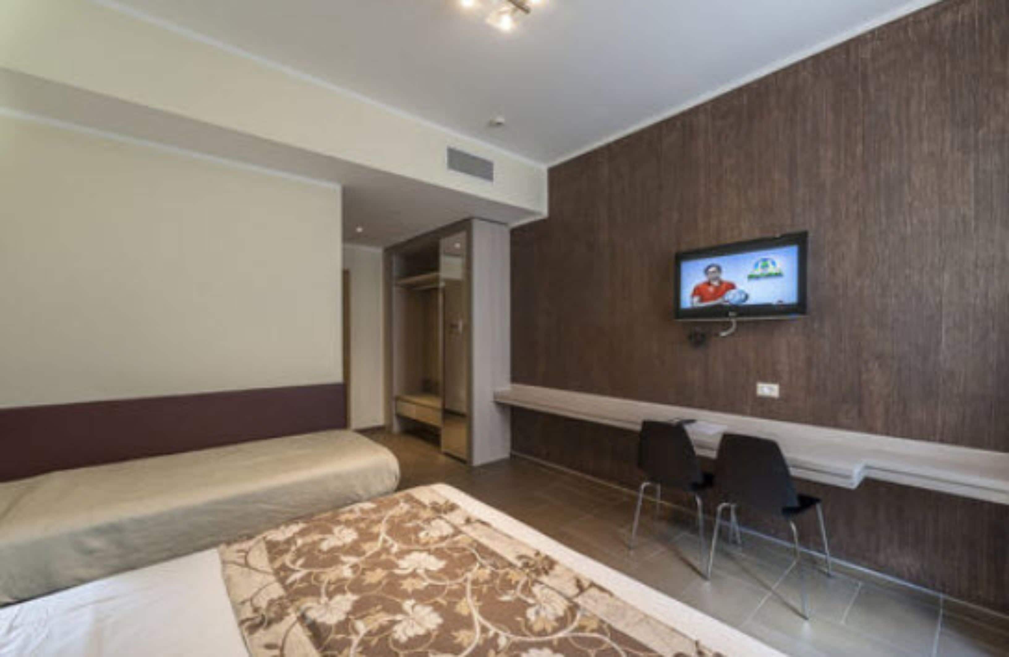 A hotel room with a bed, a desk with chairs, a television mounted on the wall, and a wardrobe.