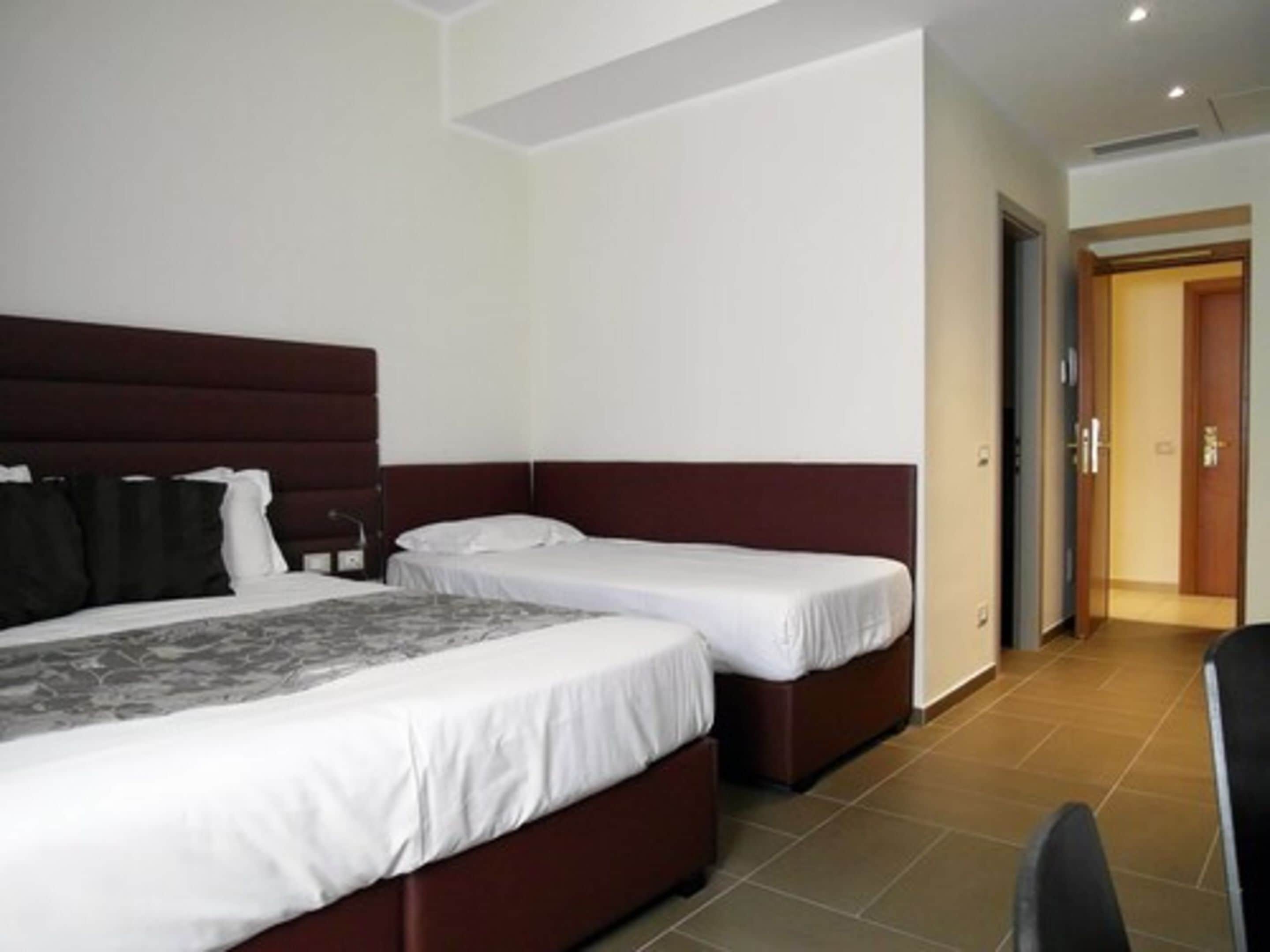 A hotel room with two beds, a large bed with a dark headboard and a smaller bed with a white headboard, both with white bedding.
