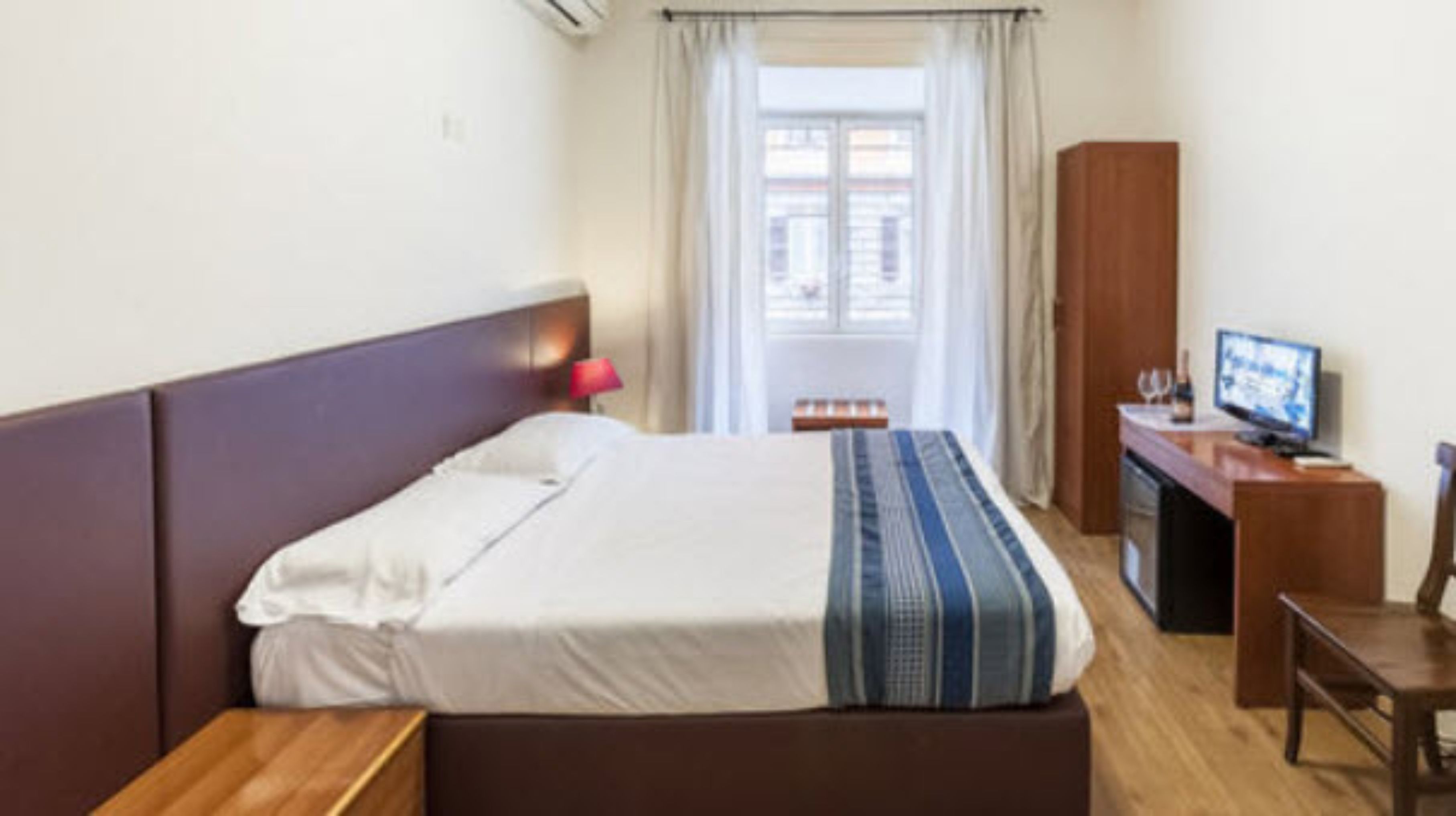Habitación de hotel con cama, escritorio, televisor y ventana con cortinas.