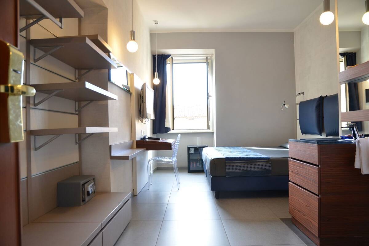 A compact hotel room with a bed, a desk, a chair, and built-in shelving.