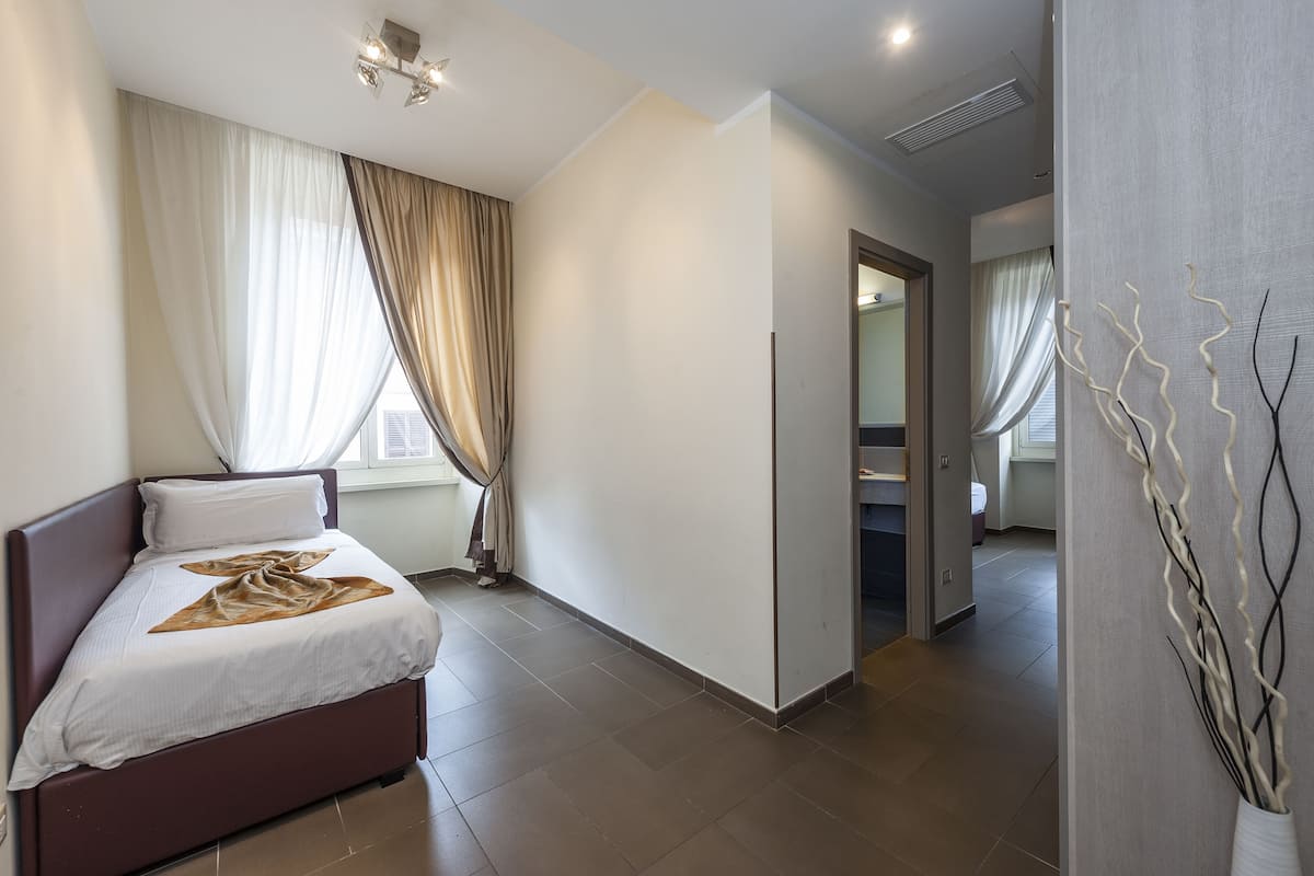 A modern hotel room with a bed, a large window with curtains, a hallway, and a bathroom visible through an open door.