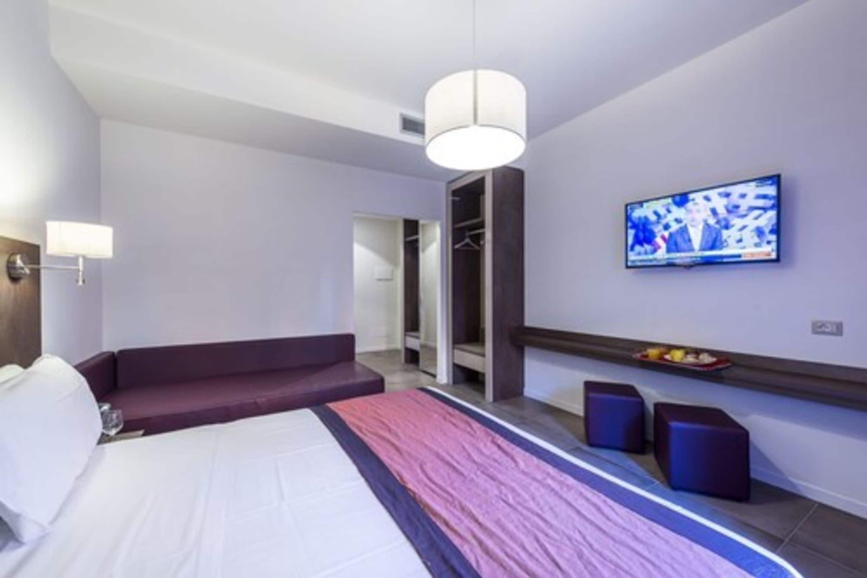 A modern hotel room with a bed, a seating area, a television, and a wardrobe.