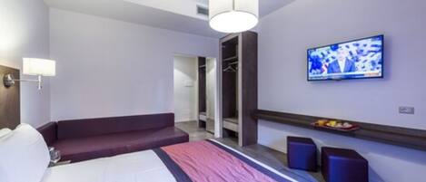 A modern hotel room with a bed, a seating area, a television, and a wardrobe.