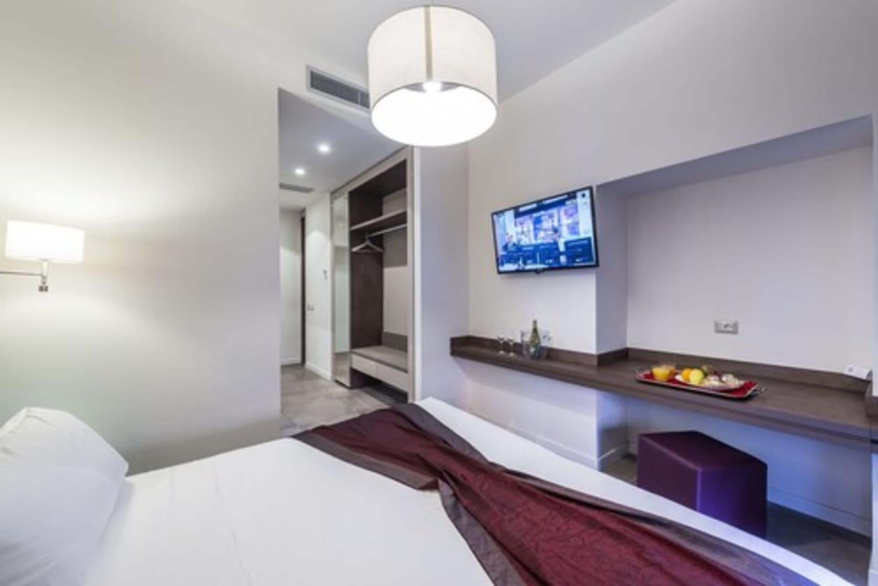 Una habitación de hotel moderna con cama, televisor montado en la pared, una pequeña zona de estar con una bandeja de frutas y un armario con espejo.