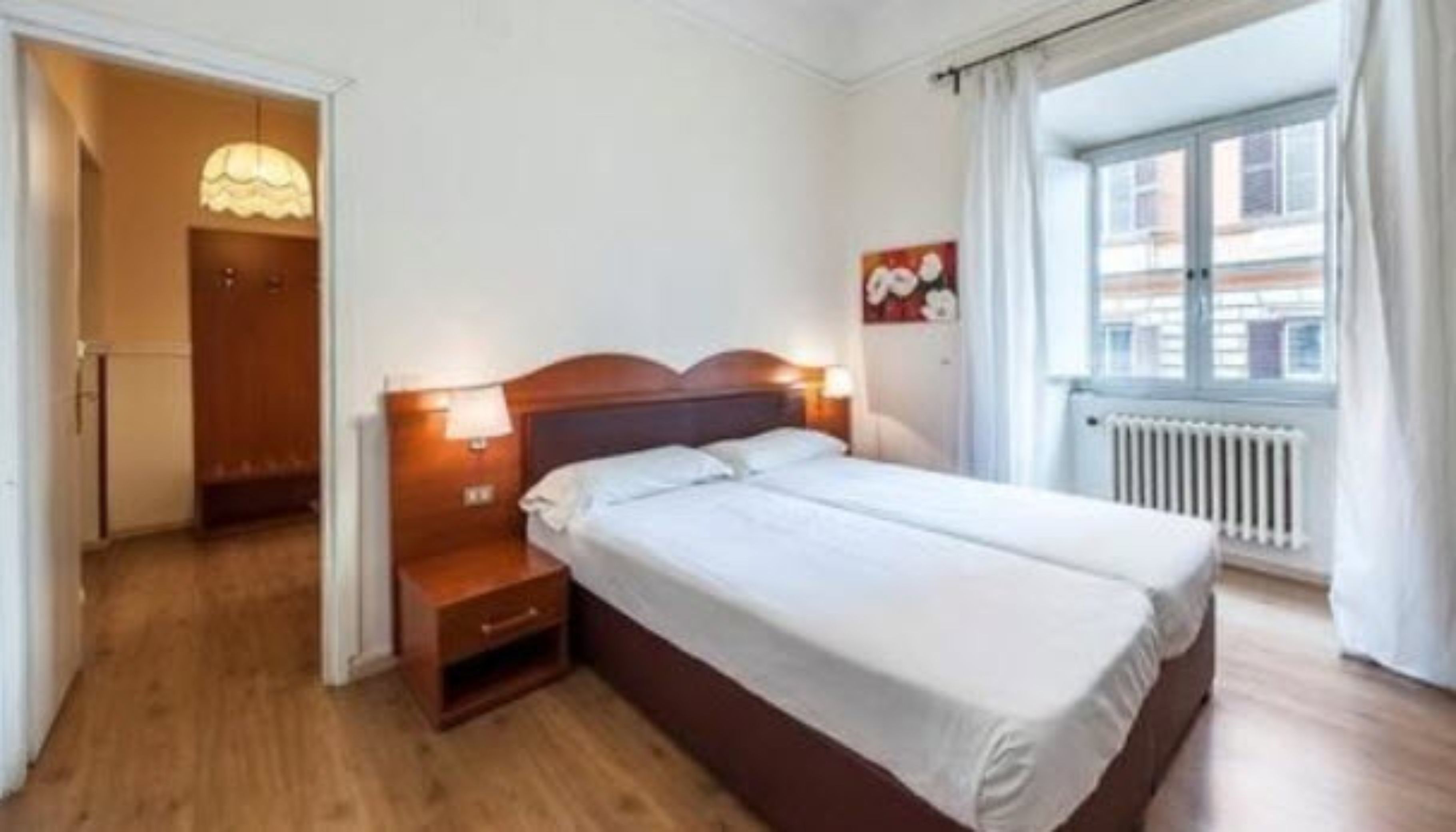 A double bed with a wooden headboard, a nightstand with a lamp, a radiator, and a window with curtains.