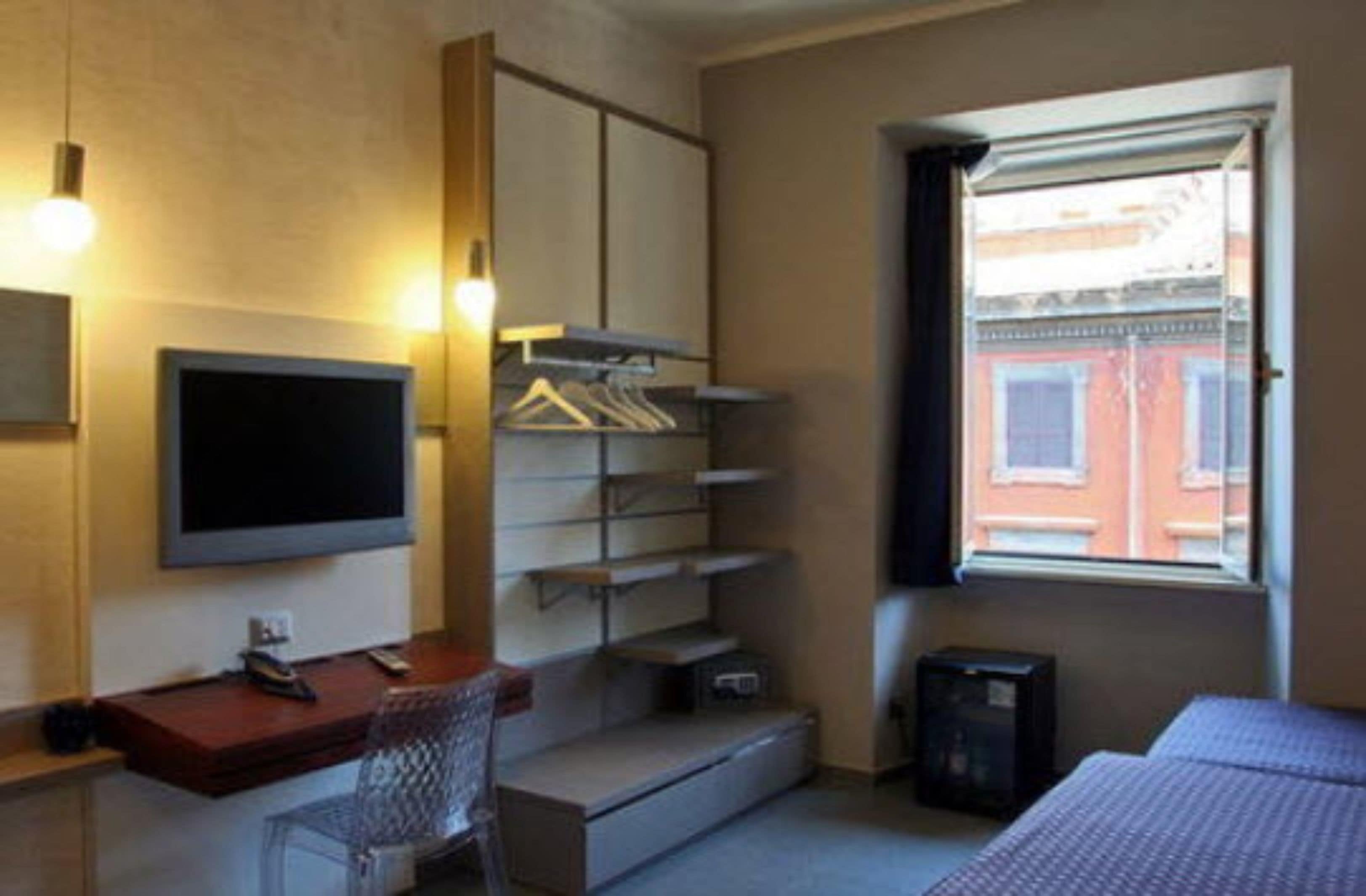 A compact hotel room with a bed, a desk with a chair, a TV, a wardrobe with shelves, and a window with a view of buildings.
