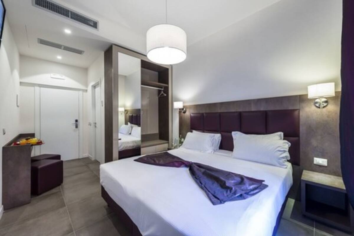 A modern hotel room with a large bed, a wardrobe, a desk, and a mirror.