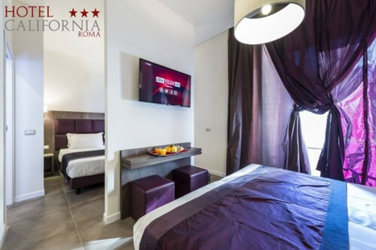A hotel room with a bed, a TV mounted on the wall, a small seating area with a tray of fruit, and a window with curtains.