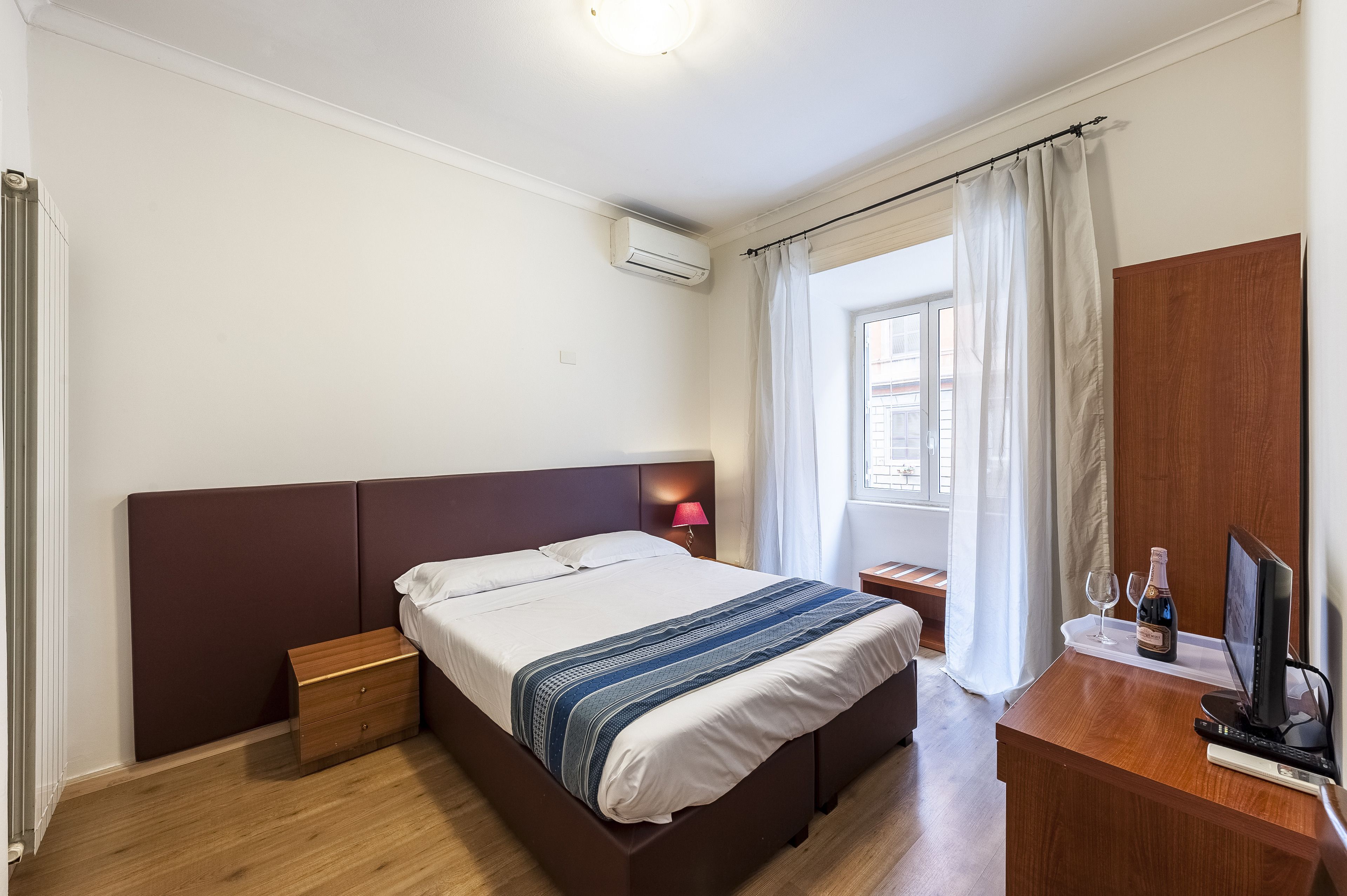 A hotel room with a bed, bedside table, lamp, TV, and a window with curtains.