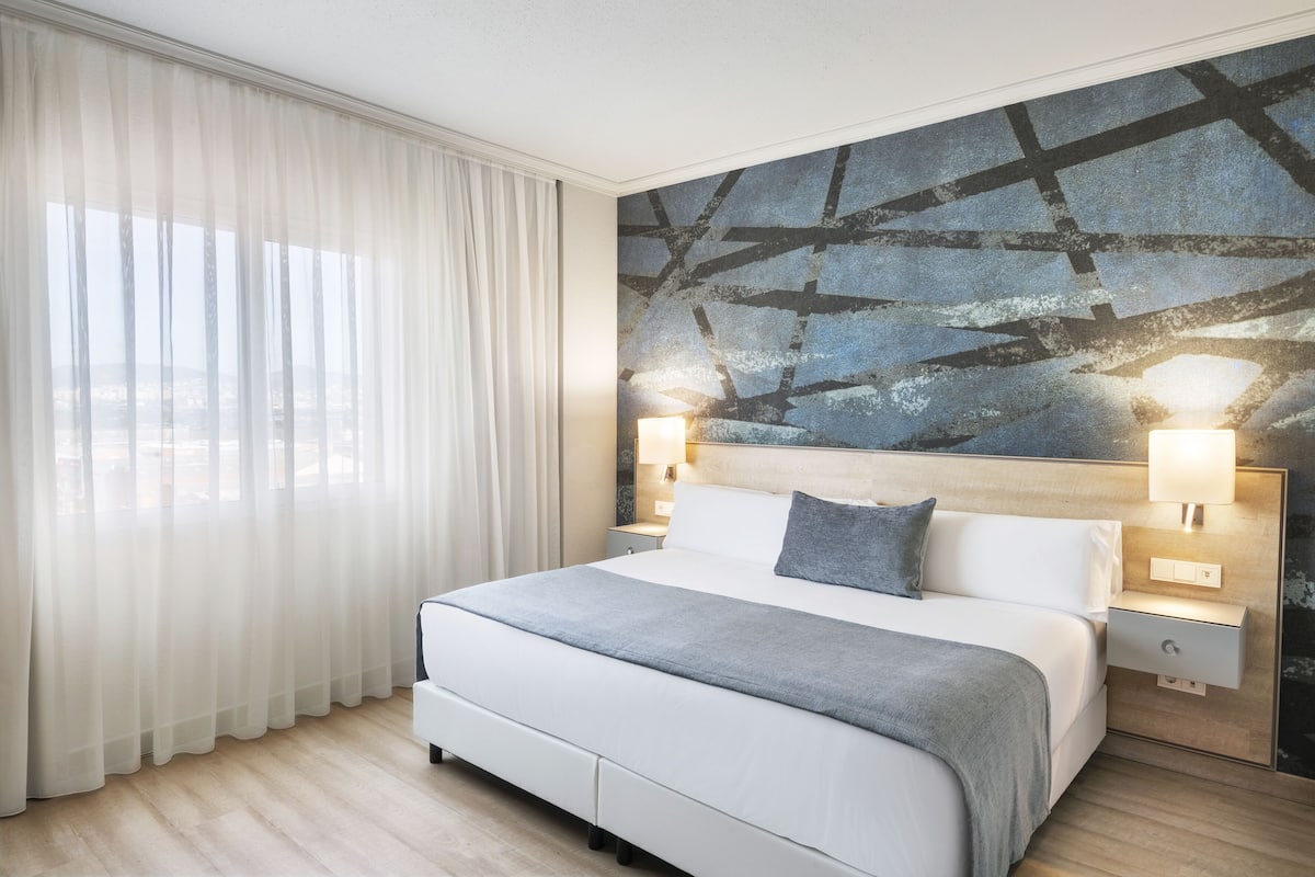 Ett modernt hotellrum med en stor säng, vita gardiner, utsikt över staden och ett unikt, abstrakt konstverk på väggen.
