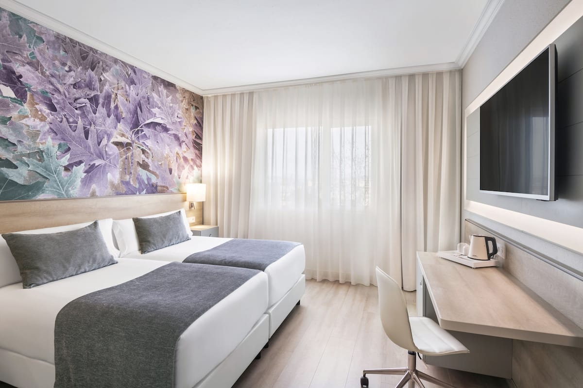 Ett hotellrum med två sängar, ett skrivbord, en stol, en TV och ett stort fönster med gardiner.