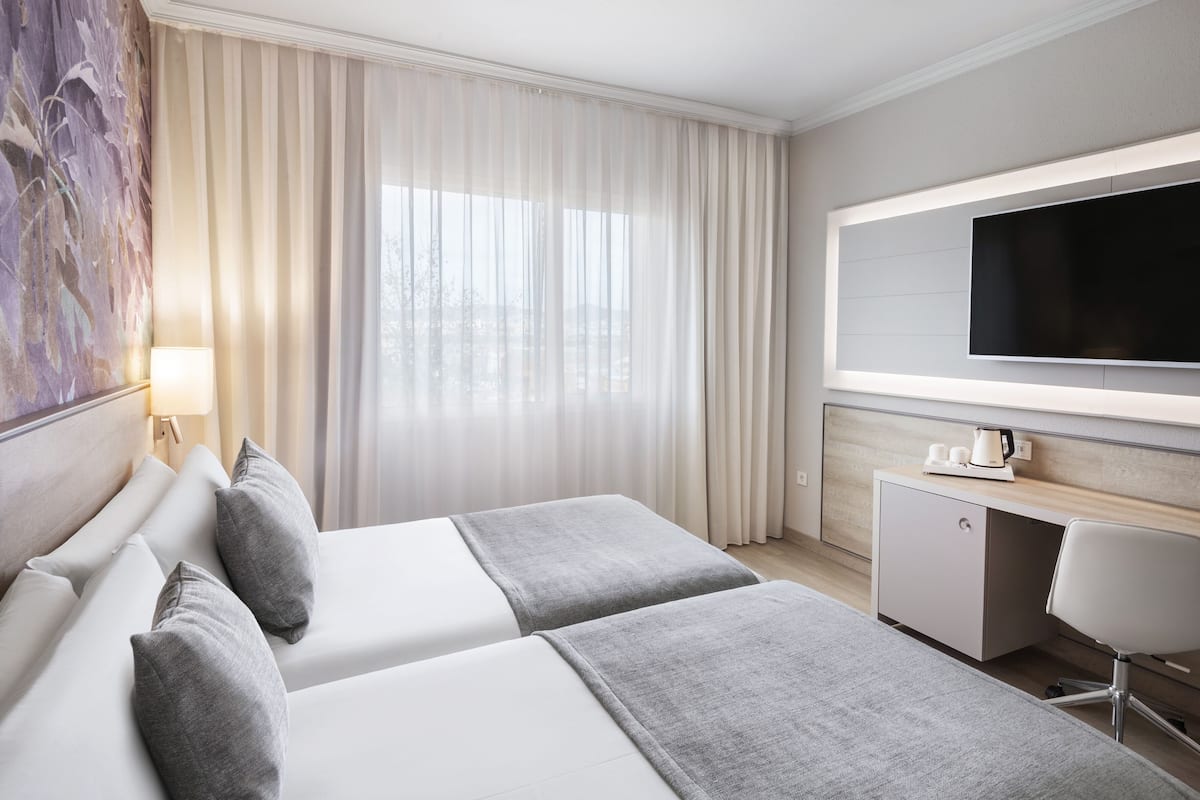 Ett hotellrum med två sängar, ett skrivbord, en platt-TV och ett stort fönster med tunna, genomskinliga gardiner.