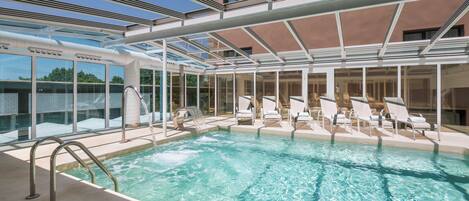 A pool area with a glass-enclosed roof, lounge chairs, and a clear view of the sky.
