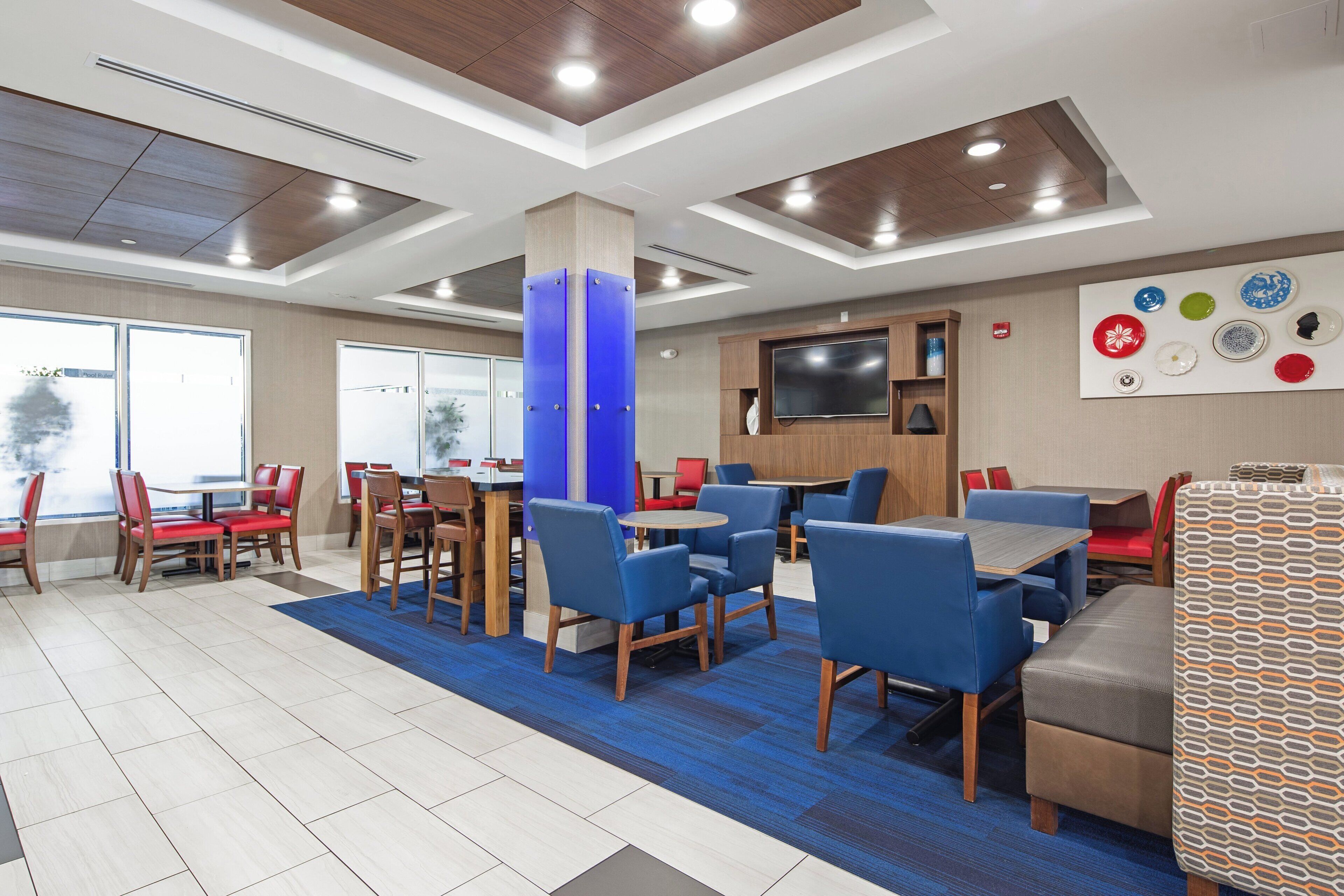 A modern dining area with multiple tables and chairs, a flat-screen TV mounted on the wall, and a blue carpet.