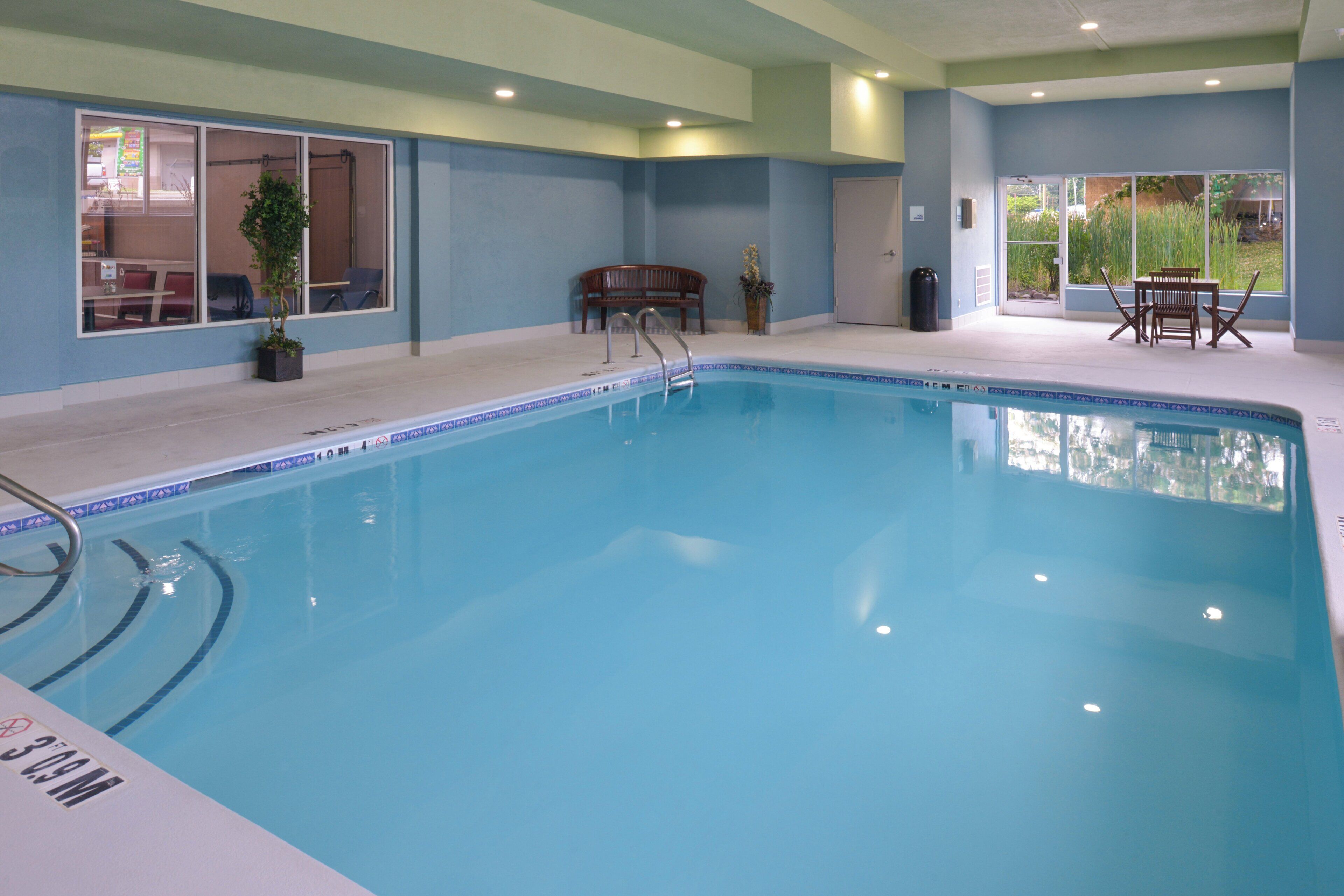 An indoor swimming pool with a tiled edge, a bench, a table with chairs, and a potted plant.