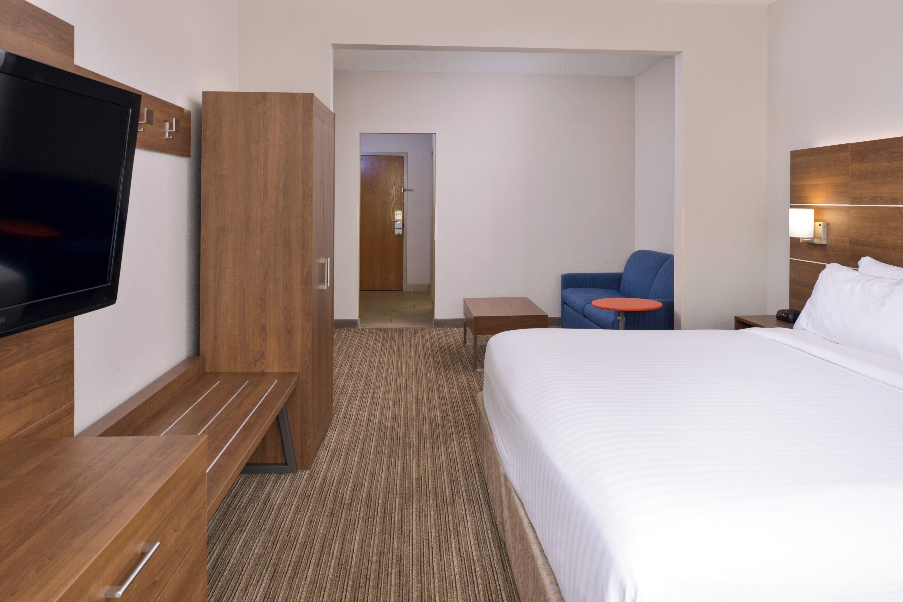 Habitación de hotel con cama, televisión, zona de estar con una silla y una mesita, y un armario de madera.
