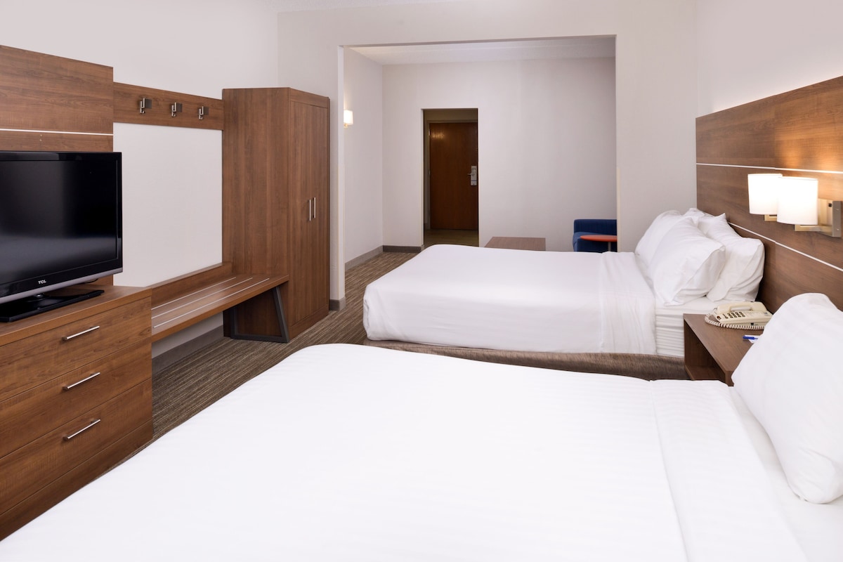 Une chambre d’hôtel avec un grand lit, un lit plus petit, un bureau avec une télévision, une armoire et une table de chevet avec une lampe.