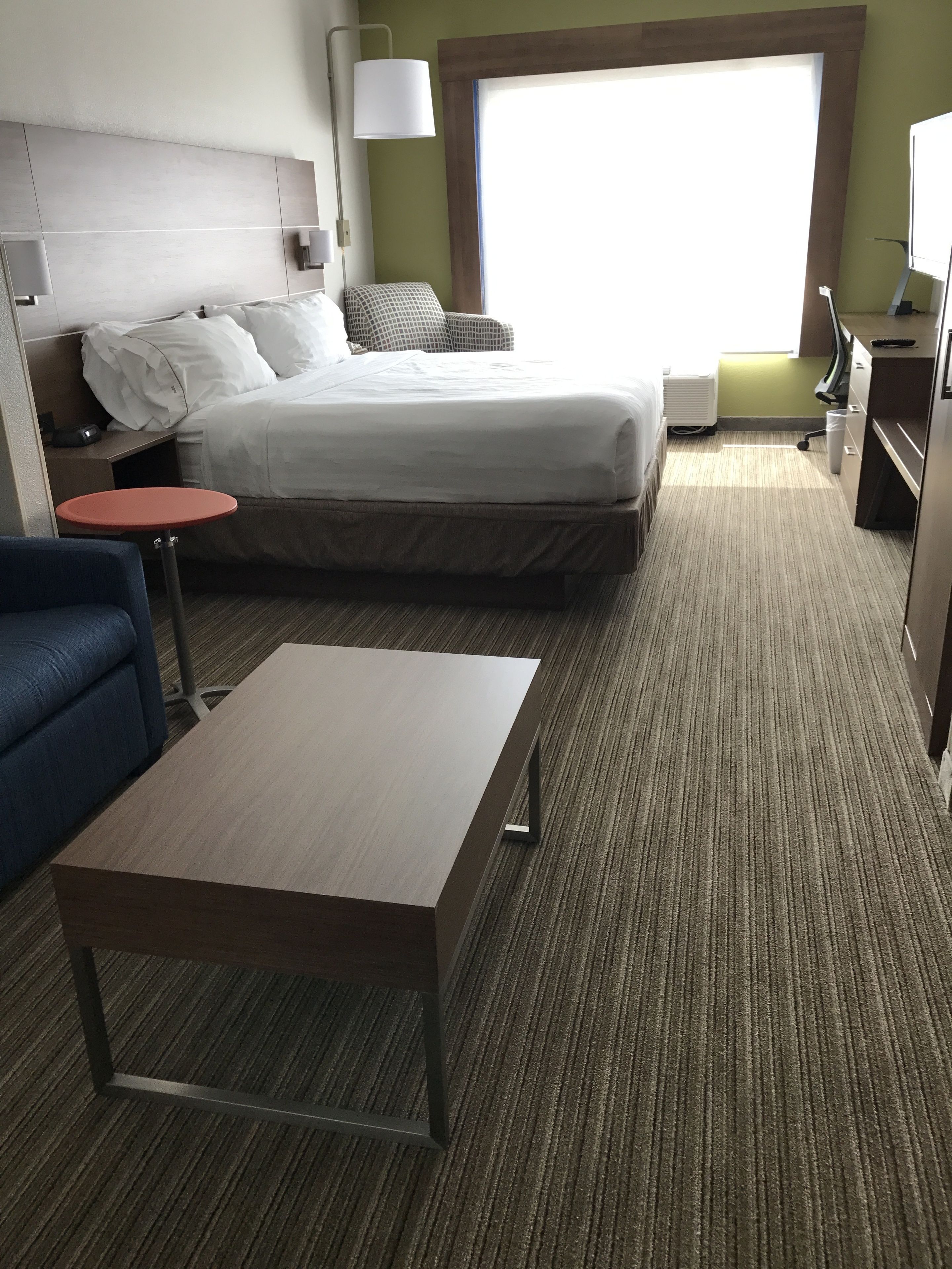A hotel room with a bed, a desk, a chair, a small table, and a lamp.