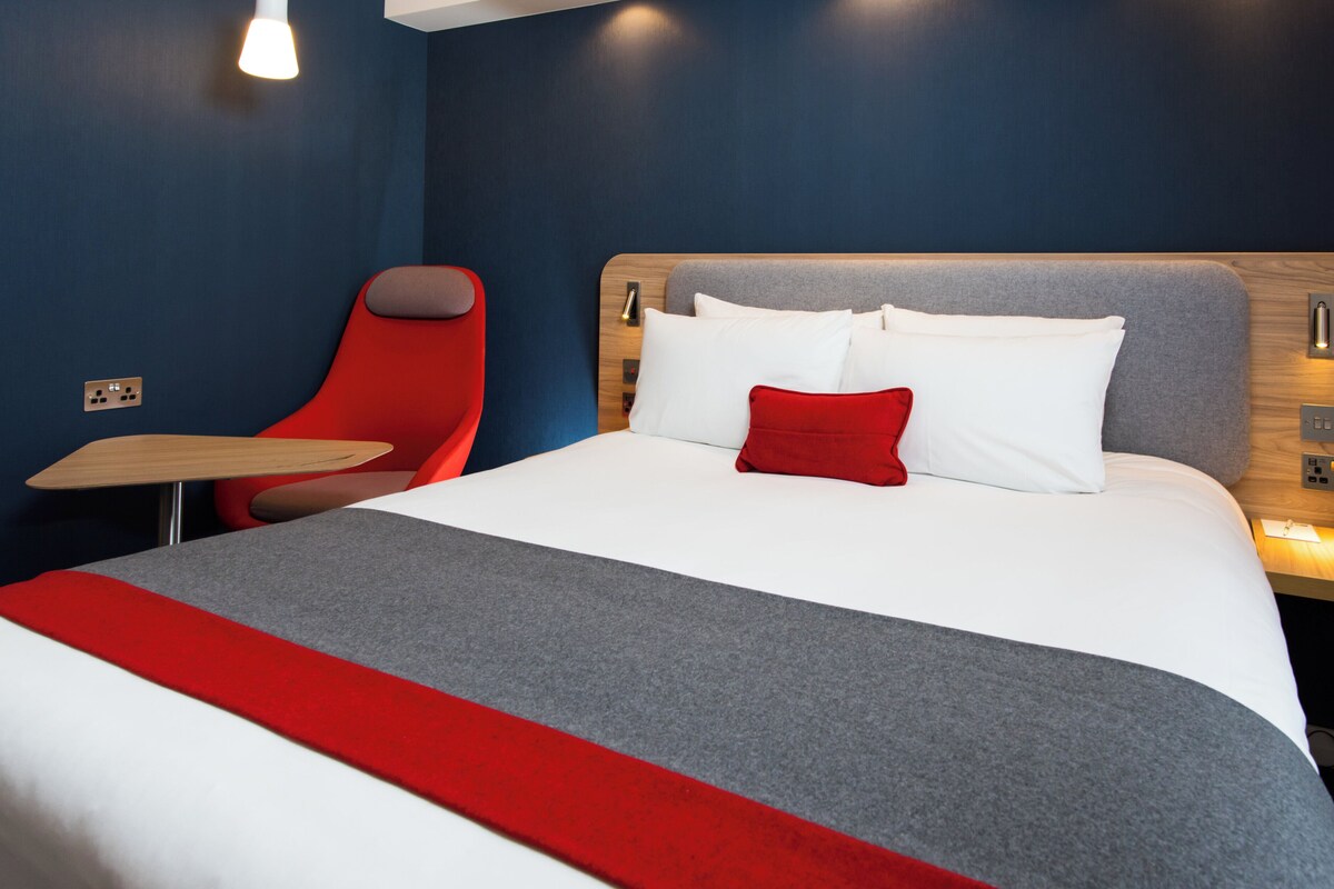 A hotel room with a bed, a red chair, a small table, and a nightstand with a lamp.