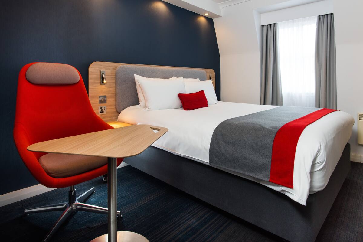 A modern hotel room with a large bed, a red chair, a wooden table, and a view of the city through the window.