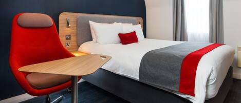 A modern hotel room with a large bed, a red chair, a wooden table, and a view of the city through the window.