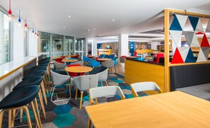 A modern cafe with round tables and colorful chairs, a counter with a person behind it, and a geometric-patterned partition. - Holiday Inn Express London Hammersmith by IHG (London)