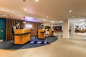 A hotel reception area with wooden reception desks, a digital display, and a seating area with tables and chairs.