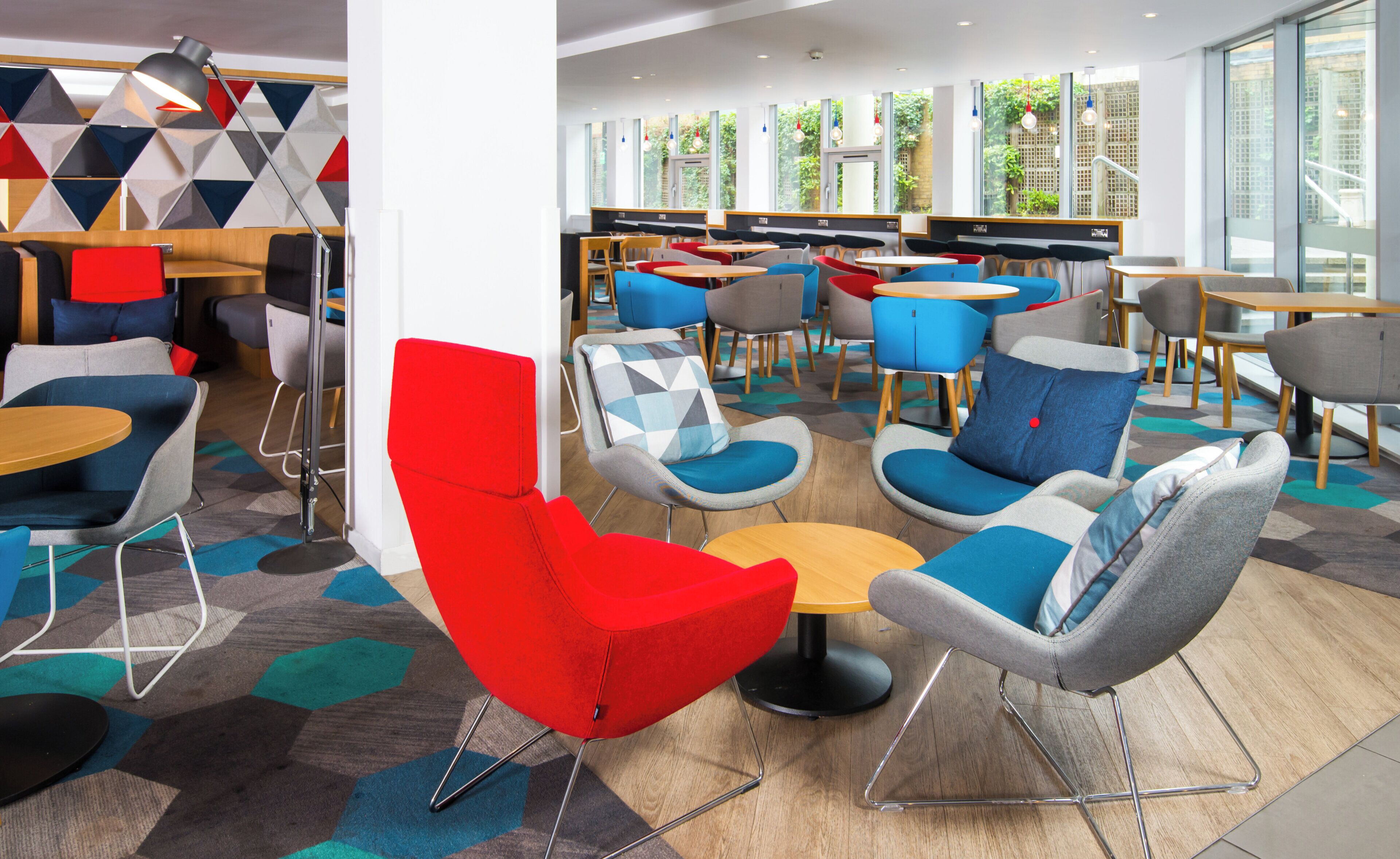 Un área de descanso moderna con varias opciones de asiento, incluyendo sillones rojos y grises, mesas y una alfombra estampada.