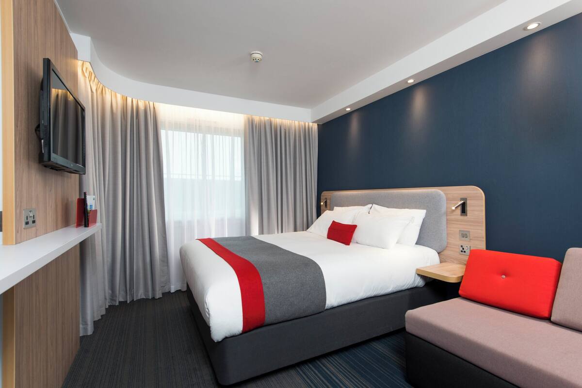 A modern hotel room with a large bed, a desk, a laptop, and a TV mounted on the wall.