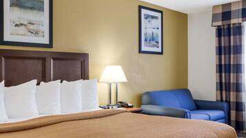 Quarto de hotel com uma cama grande, uma escrivaninha com luminária, uma poltrona azul e duas fotos emolduradas na parede.