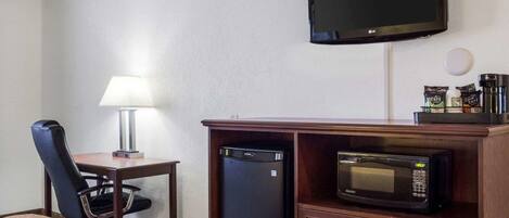 A hotel room with a bed, a desk with a lamp, a television mounted on the wall, a microwave, and a coffee machine.