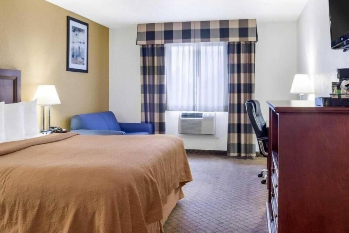 Ett hotellrum med en säng, ett skrivbord, en stol, en tv och ett fönster med gardiner.