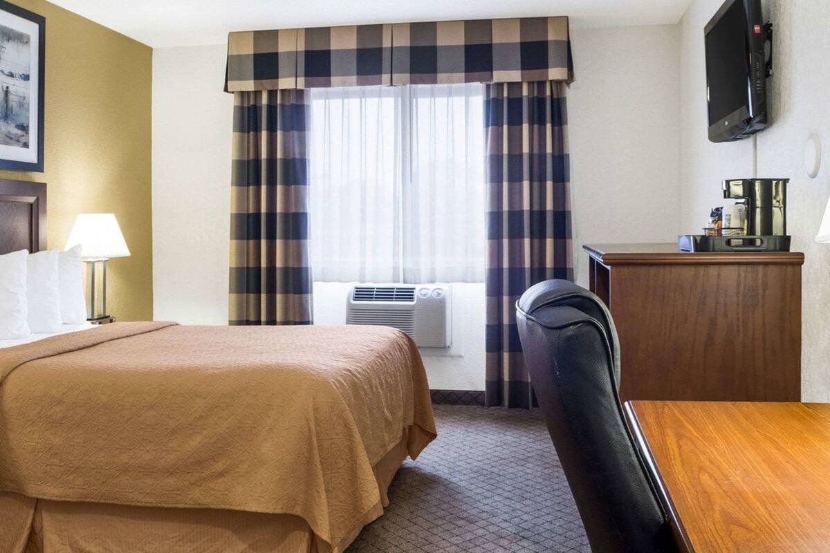 Ett hotellrum med en säng, ett skrivbord, en stol, en tv och ett fönster med gardiner.