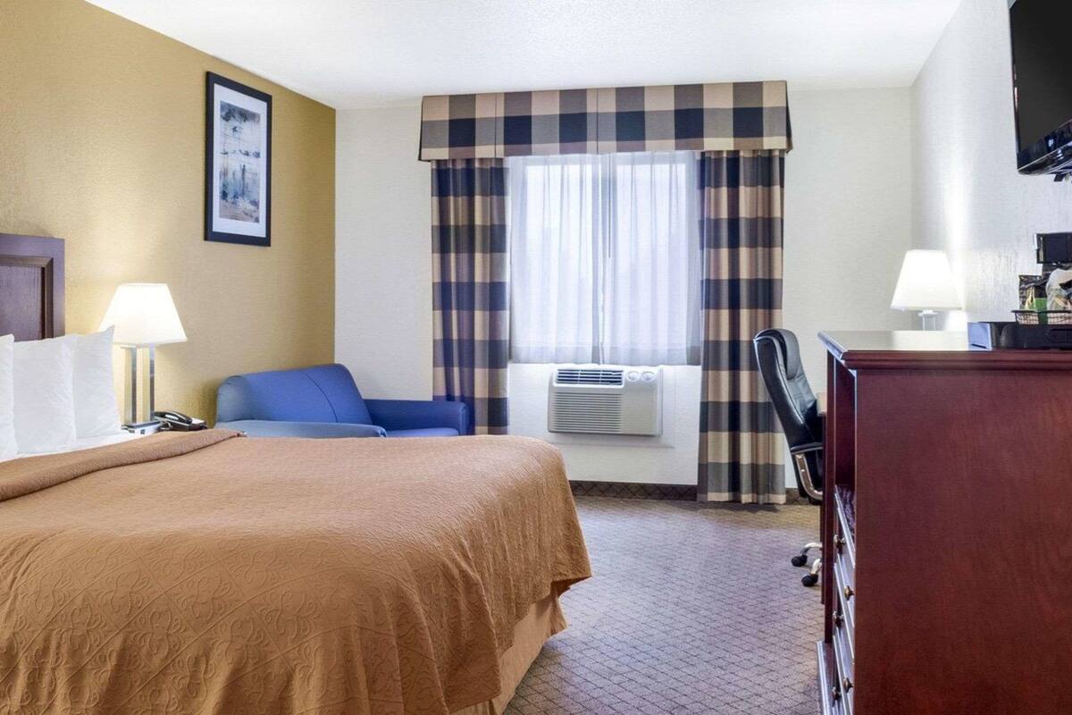 Ett hotellrum med en säng, ett skrivbord, en stol, en lampa, en luftkonditionering och ett fönster med gardiner.