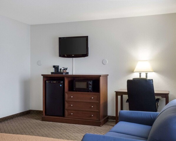 Habitación de hotel con una televisión de pantalla plana montada en la pared, un mueble de madera con microondas, cafetera y una mesita con lámpara.