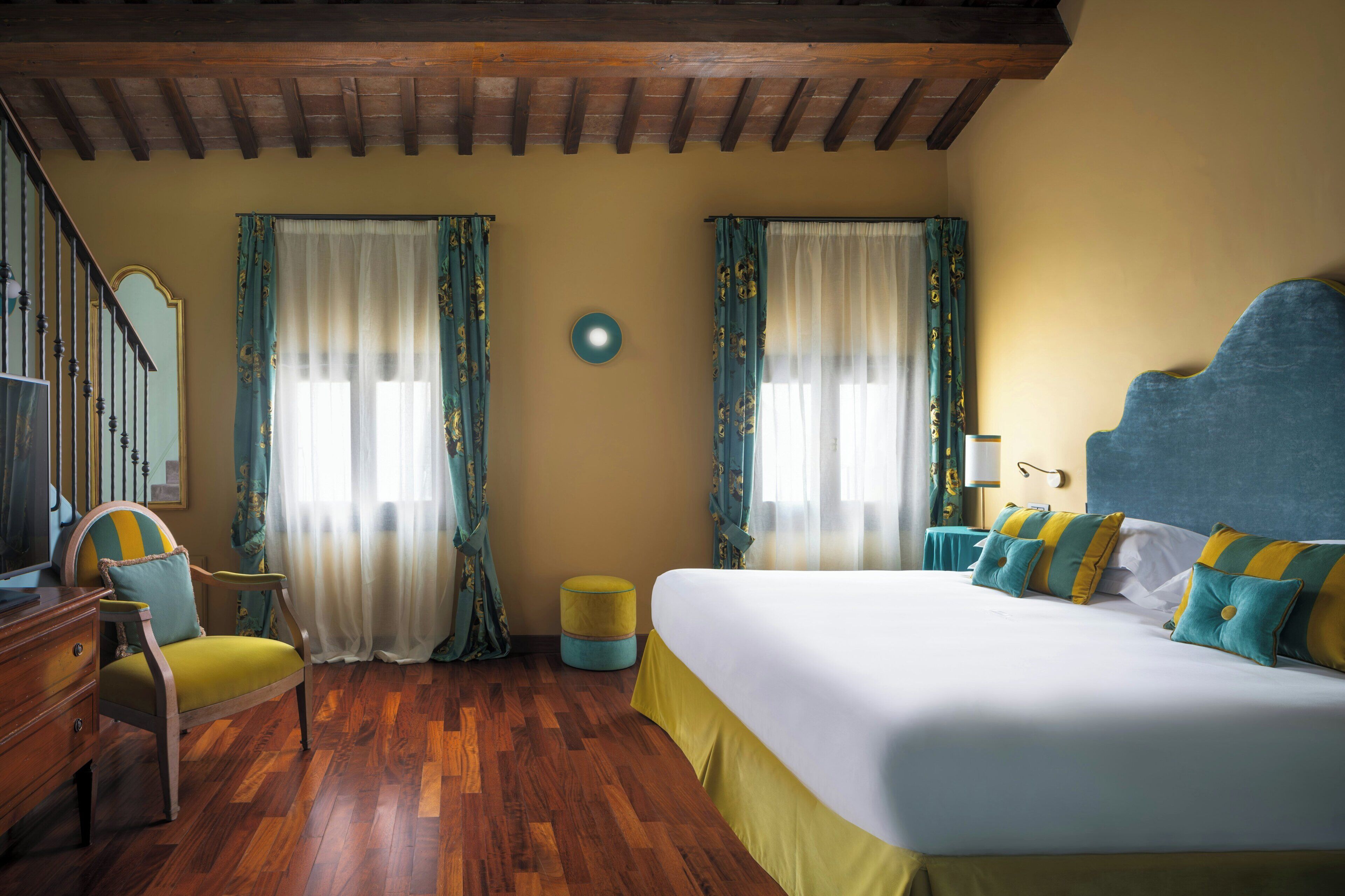 Habitación de hotel con techo de madera, cama con ropa de cama amarilla y azul, una silla, un reposapiés pequeño y redondo, y un espejo en la pared.