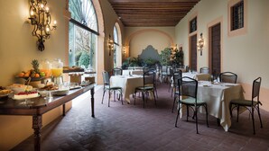 A dining area with tables and chairs, a buffet with fruits and beverages, and a view of the outdoors through large windows.