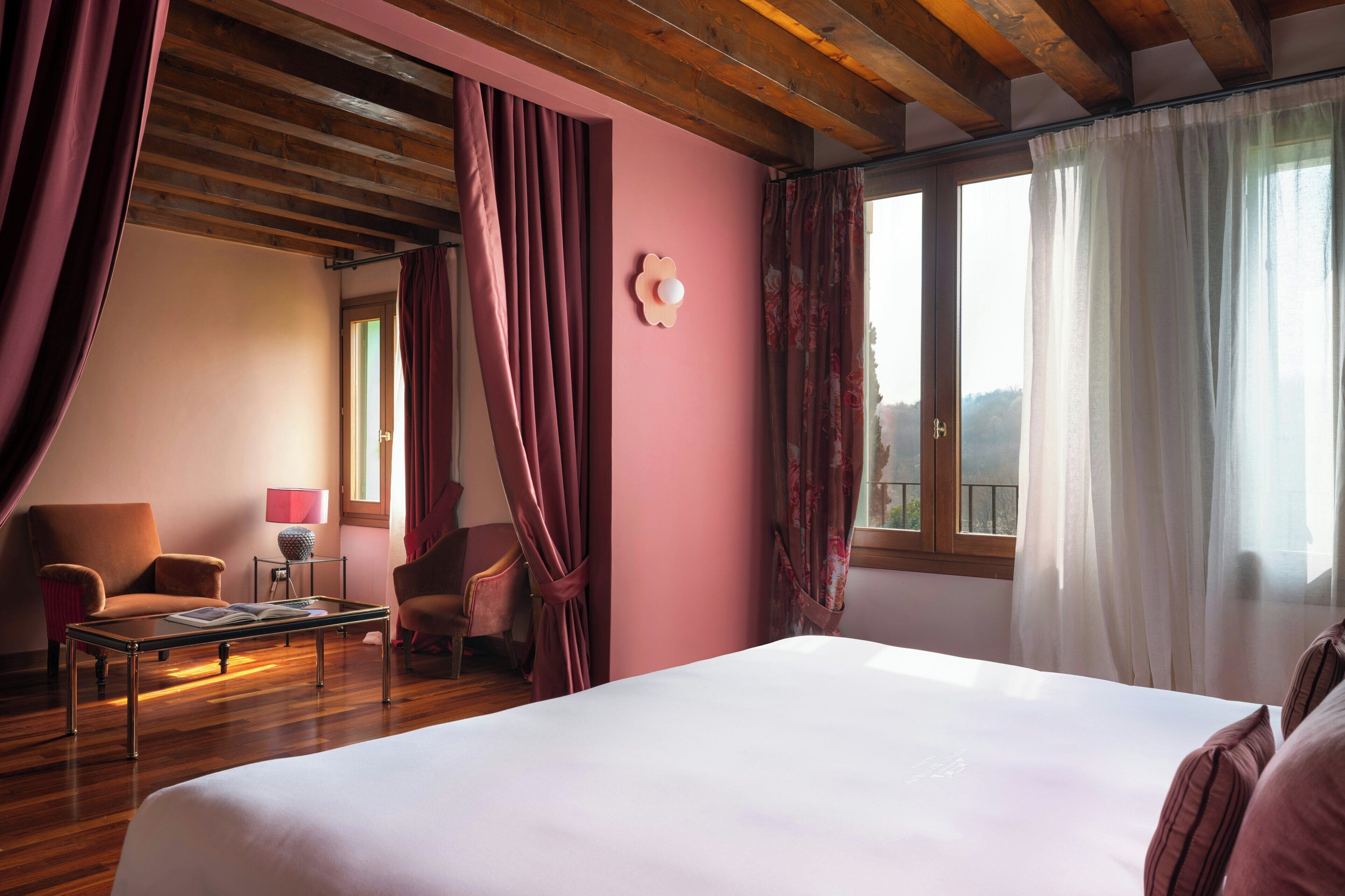 Habitación de hotel con una cama grande, vigas de madera en el techo, una ventana con cortinas, una mesita con una lámpara y dos sillas.