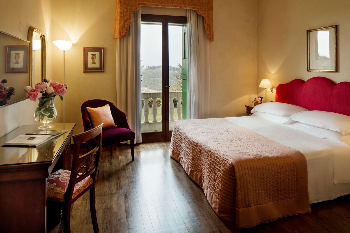Habitación de hotel con cama, escritorio con silla, un florero con flores y vistas a través de un ventanal.