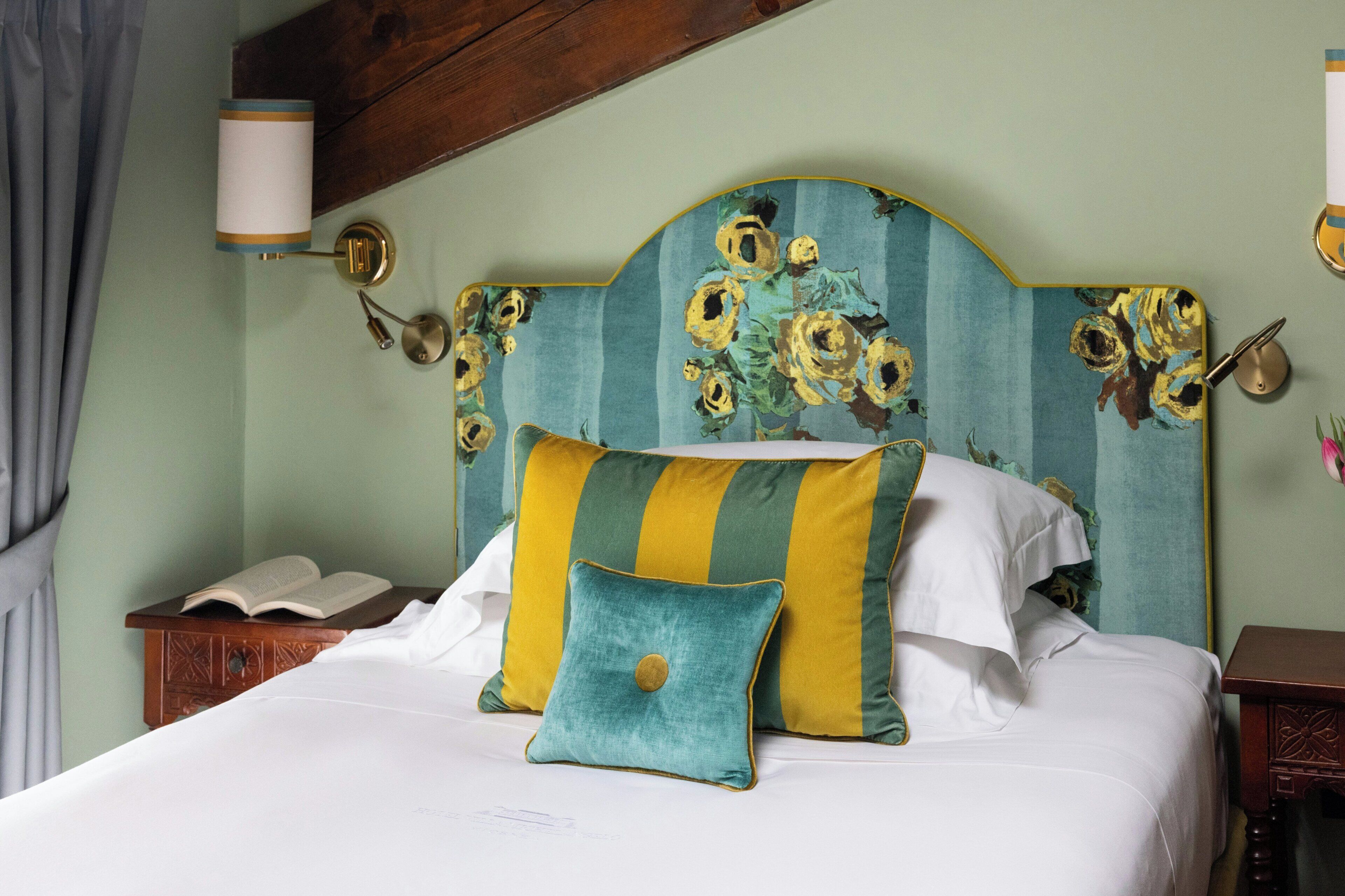 Una cama con cabecera floral, una lámpara de noche y una mesita de noche con un libro.