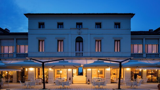 A large, elegant building with a central entrance, multiple balconies, and outdoor seating areas with umbrellas.