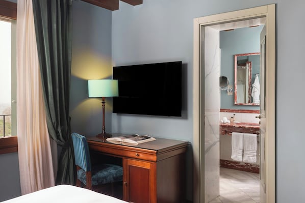 A hotel room with a bed, a desk with a lamp, a flat-screen TV, a bathroom with a mirror and sink. - Villa Michelangelo Vicenza – Starhotels Collezione (Arcugnano)
