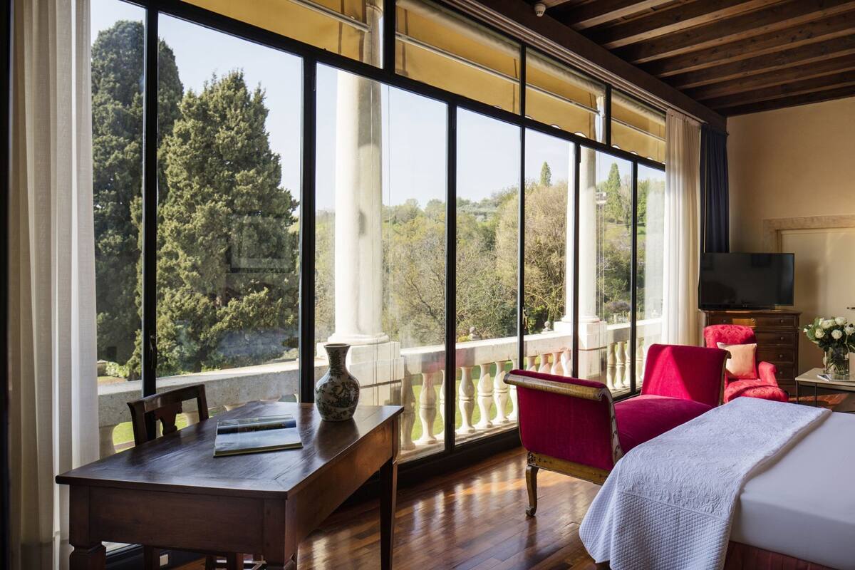 Una habitación con ventanas amplias, una mesa de madera, una silla roja, una cama y un televisor.