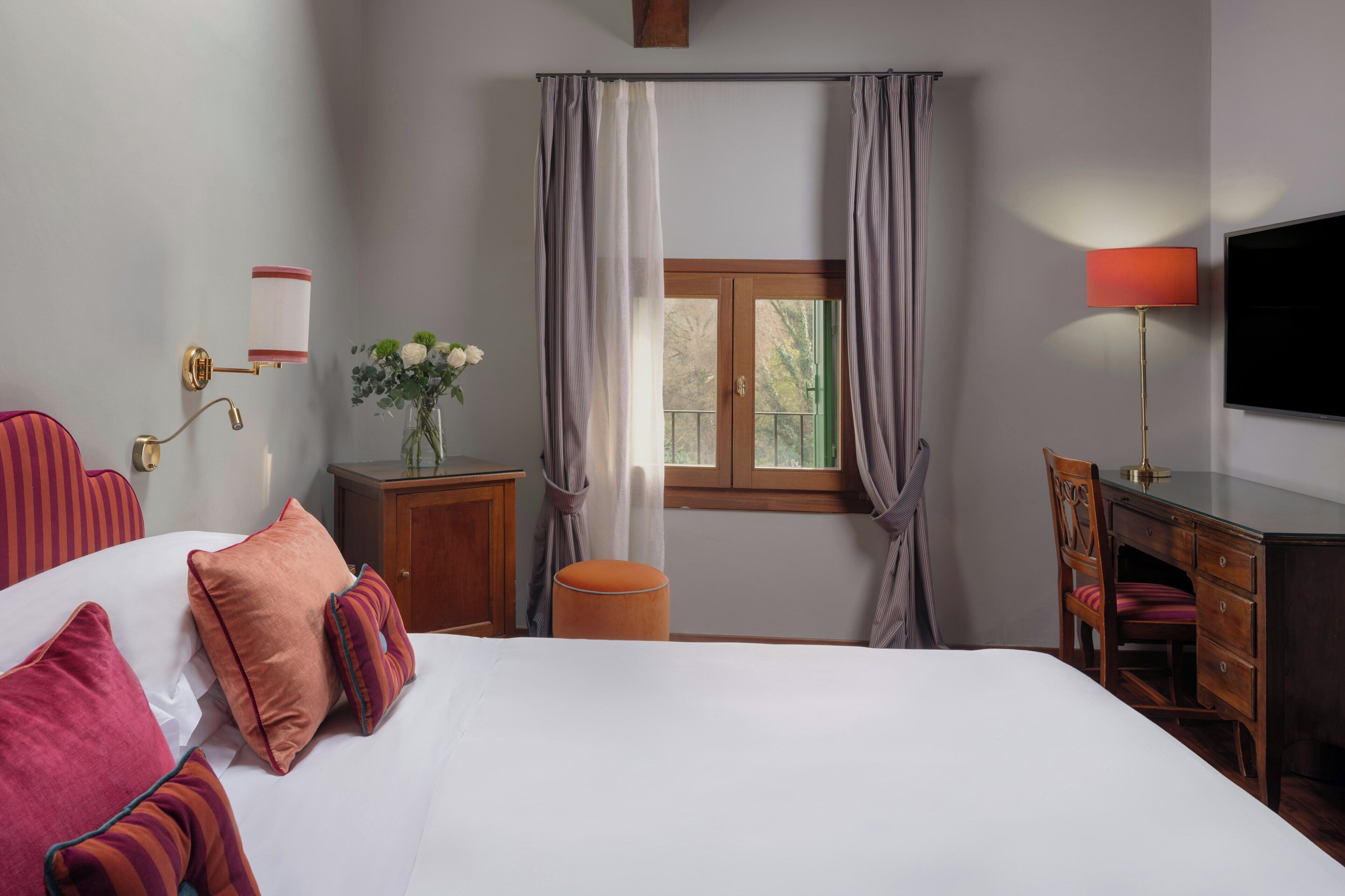Une chambre d’hôtel comprenant un lit, un bureau, une chaise, une télévision, une fenêtre avec des rideaux, une petite table avec un vase et une lampe fixée au mur.