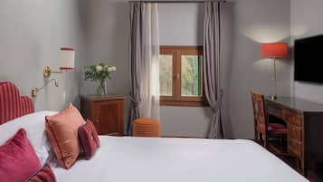Une chambre d’hôtel comprenant un lit, un bureau, une chaise, une télévision, une fenêtre avec des rideaux, une petite table avec un vase et une lampe fixée au mur.