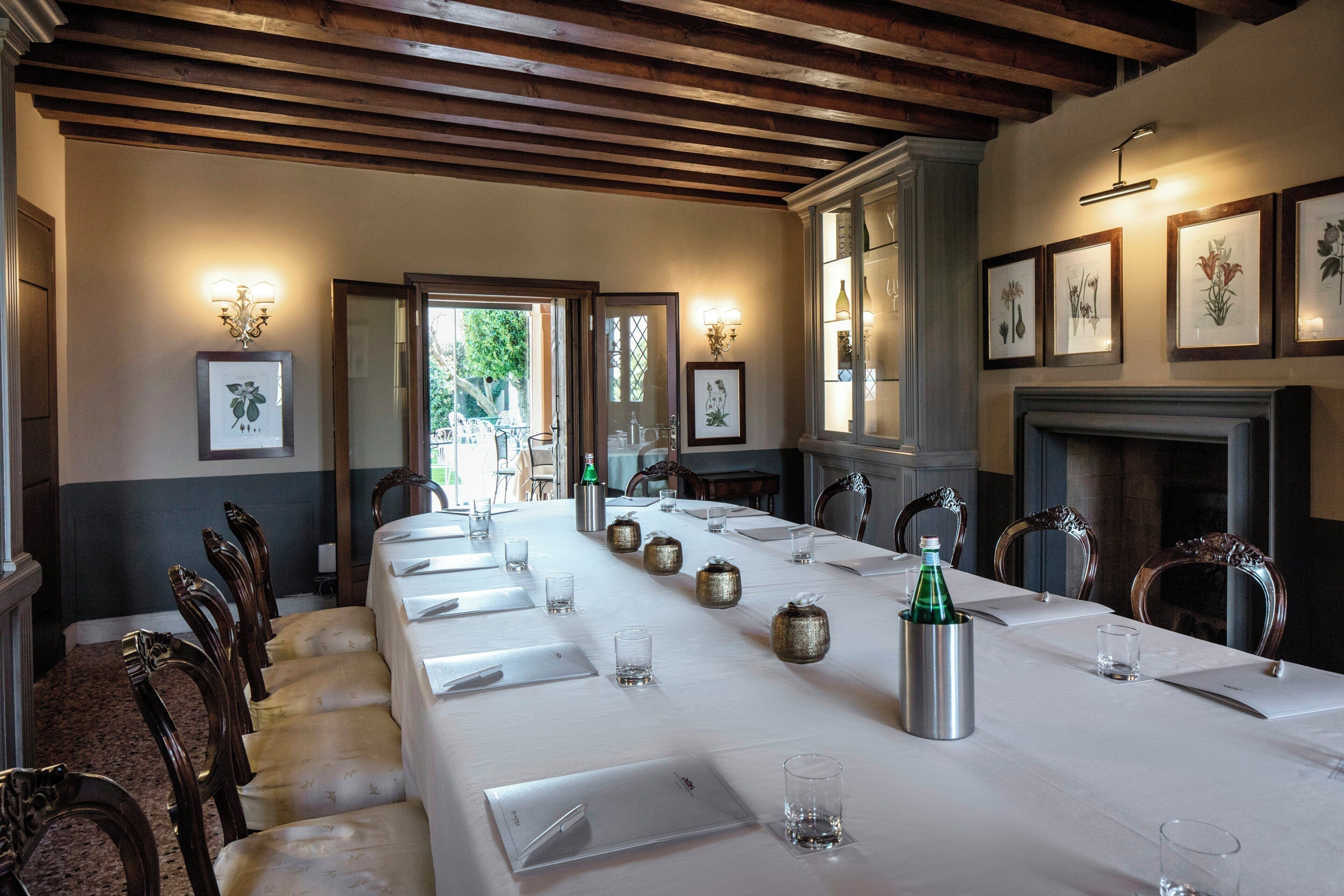 Una larga mesa de comedor dispuesta para una comida, sillas de madera, obras de arte botánicas enmarcadas en las paredes y una chimenea.