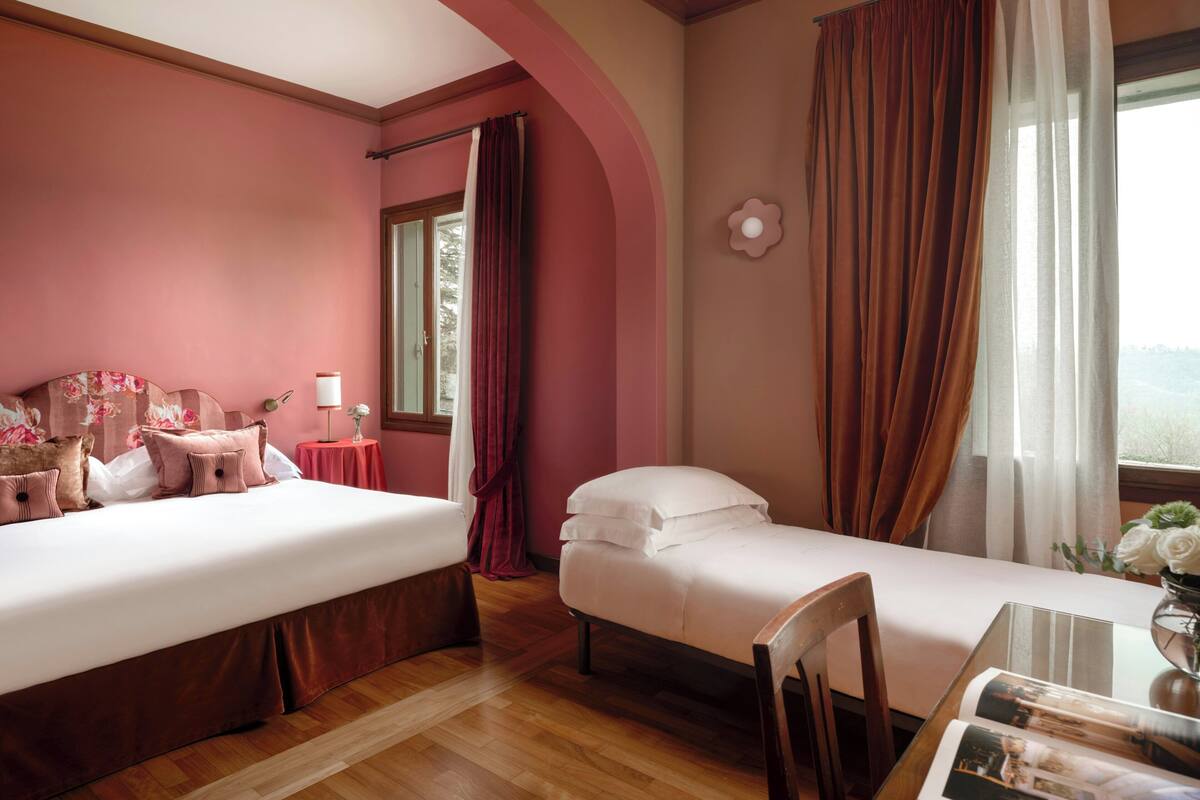 Una habitación de hotel con dos camas, una mesa de comedor y un jarrón de flores, y una ventana con cortinas.