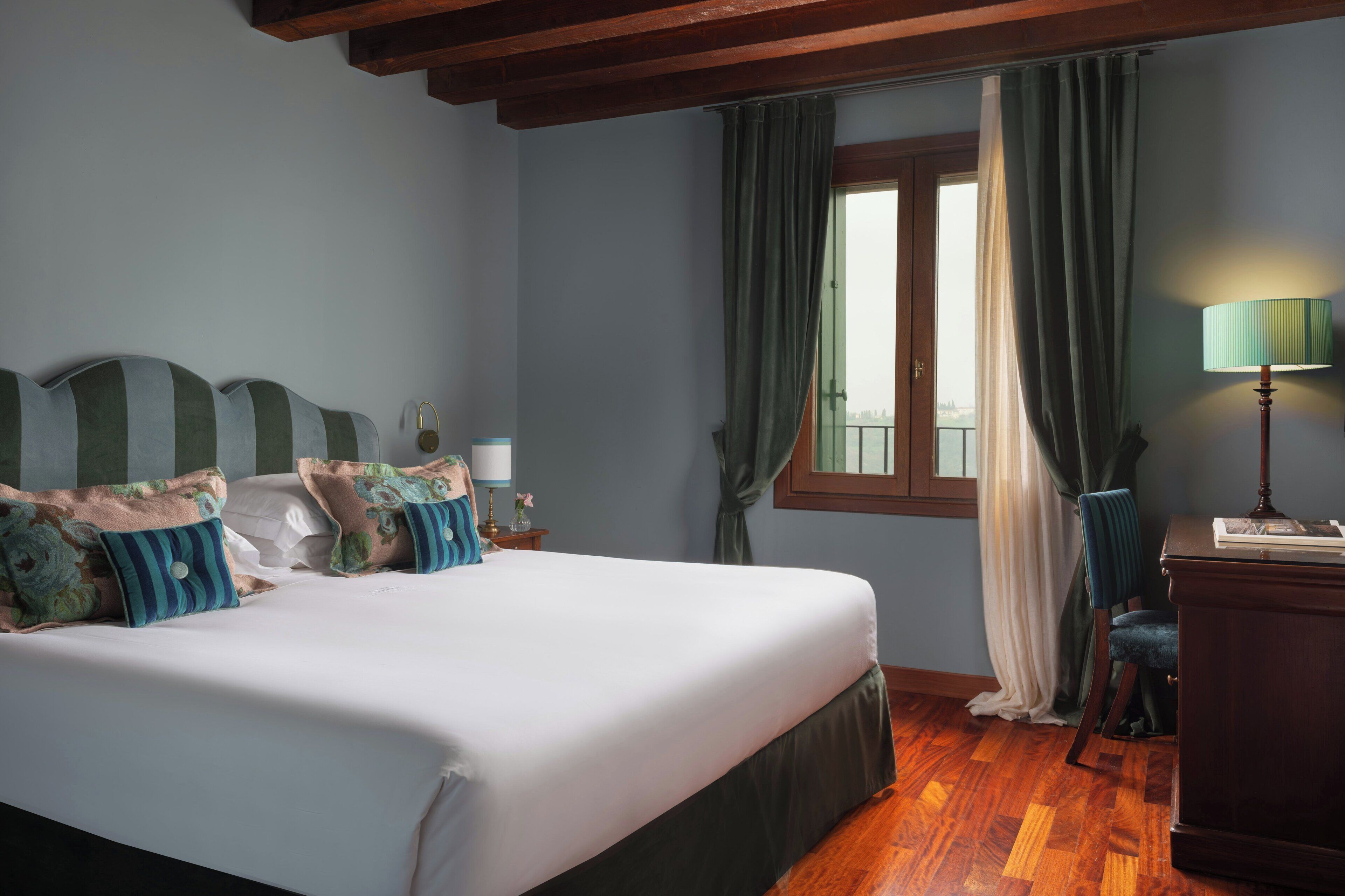 A bedroom with a large bed, a desk, a chair, a window with curtains, and a lamp.