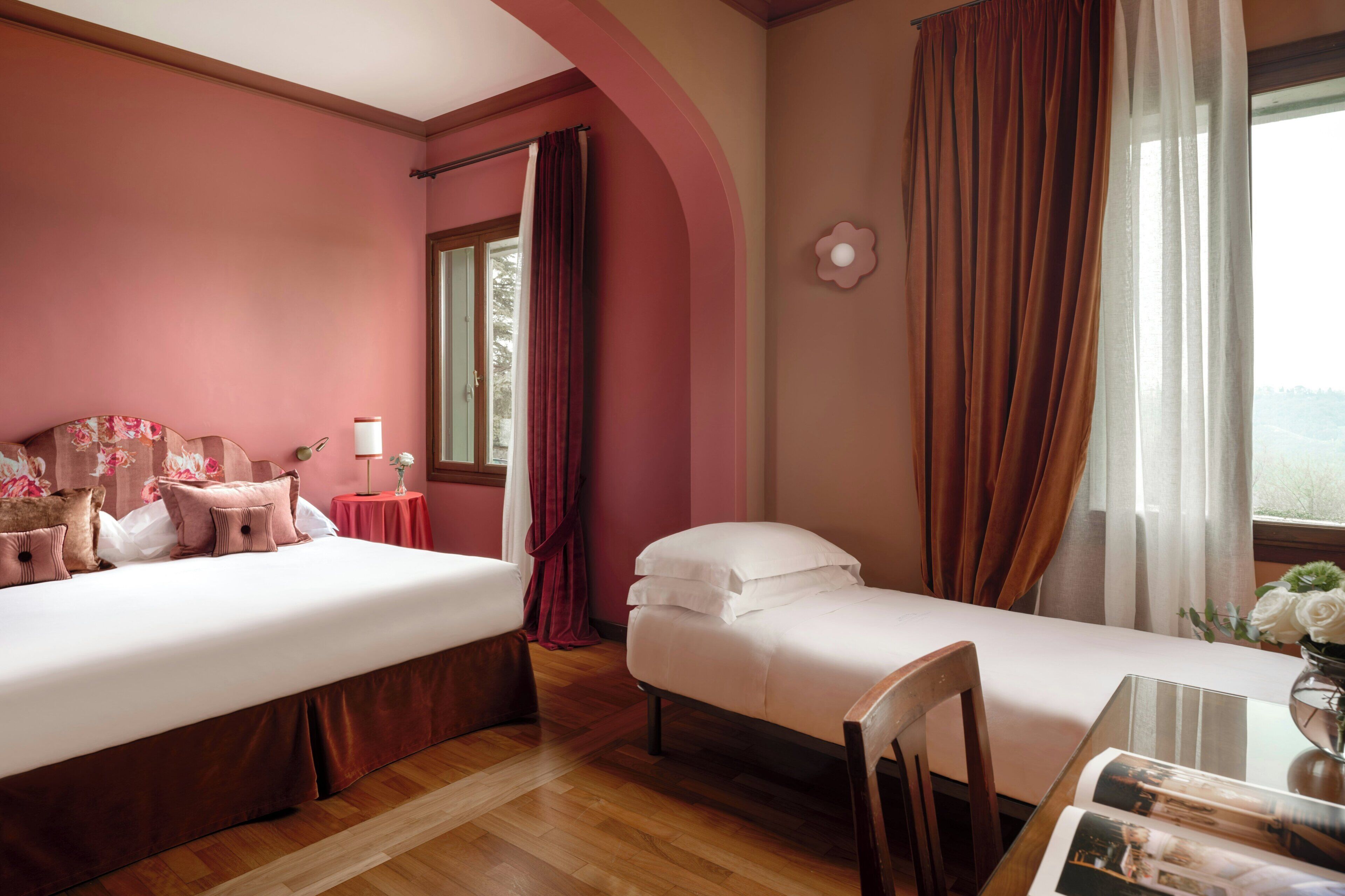 Ein Hotelzimmer mit zwei Betten, einem Esstisch mit Blumenvase und einem Fenster mit Vorhängen.